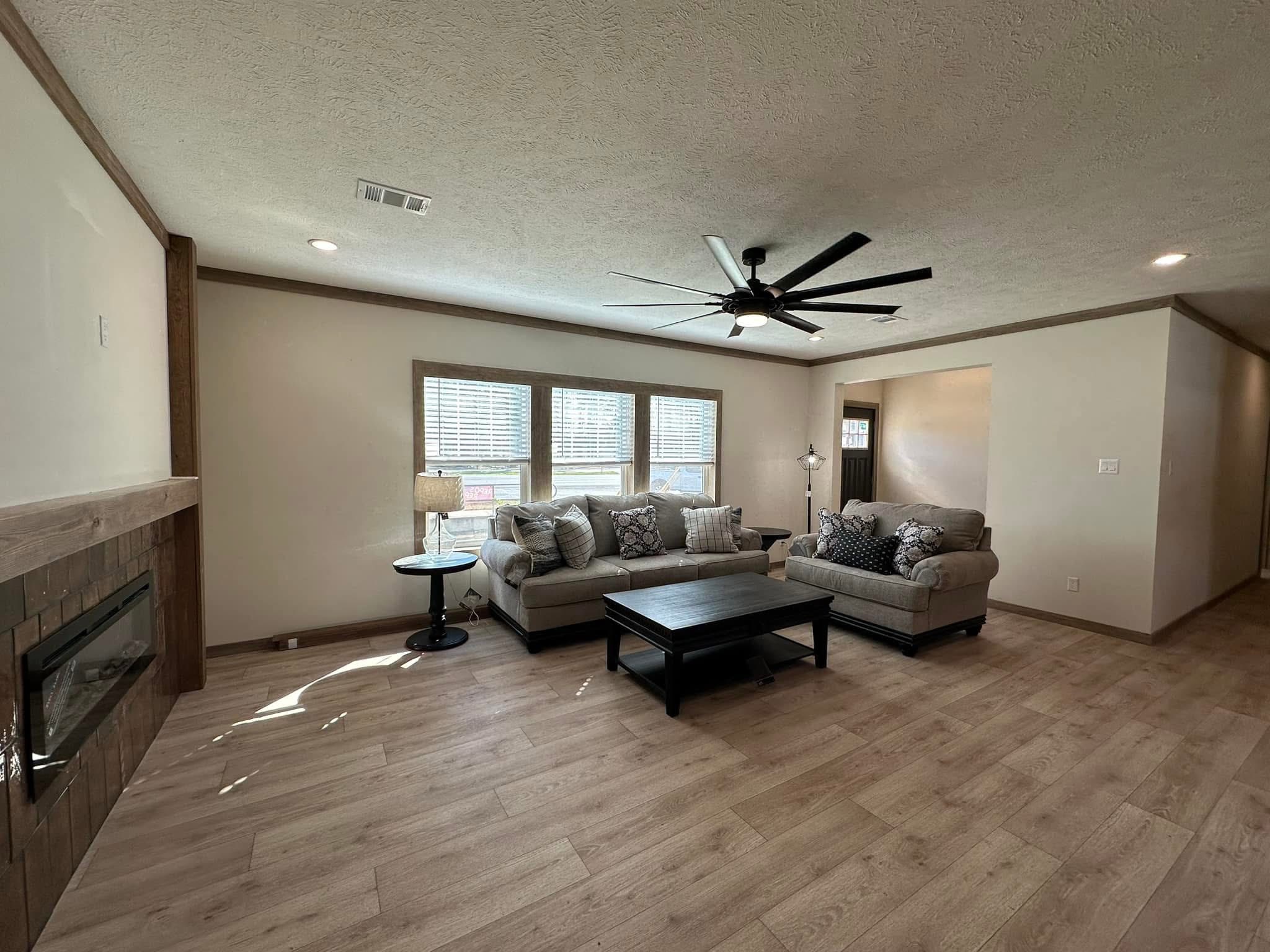 Spacious living room with light wood flooring, gray sofas adorned with patterned cushions, and a black coffee table. Large window allows natural light, illuminating a cozy, welcoming atmosphere. Ceiling fan above; modern fireplace to the left.