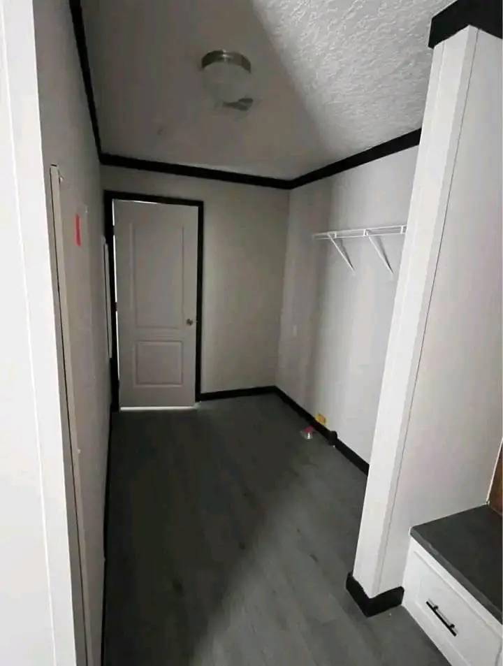 A narrow, dimly lit hallway with gray floors and white walls. A closed door at the end, a white shelf on the right, and a ceiling light fixture.