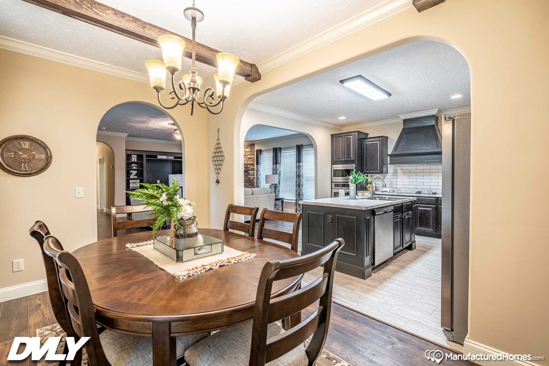 Warm dining room with a wooden table, surrounded by chairs, under an elegant chandelier. Archways lead to a modern kitchen with dark cabinetry. Cozy and inviting ambiance.