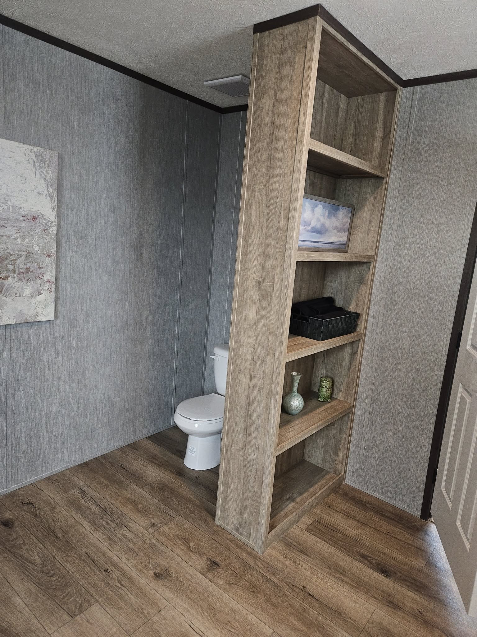 Modern bathroom with wood shelves beside a toilet. Shelves hold decor items like vases and a framed picture. Gray walls and wood flooring. Calm atmosphere.