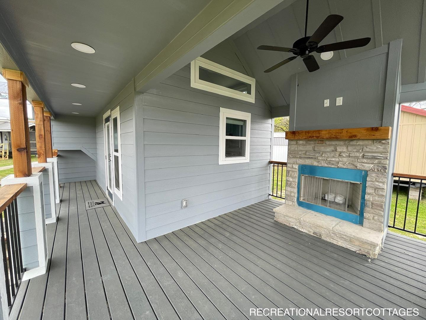 Covered patio with light gray siding and a stone fireplace featuring a blue accent. Ceiling fan overhead with recessed lighting creates a cozy vibe.