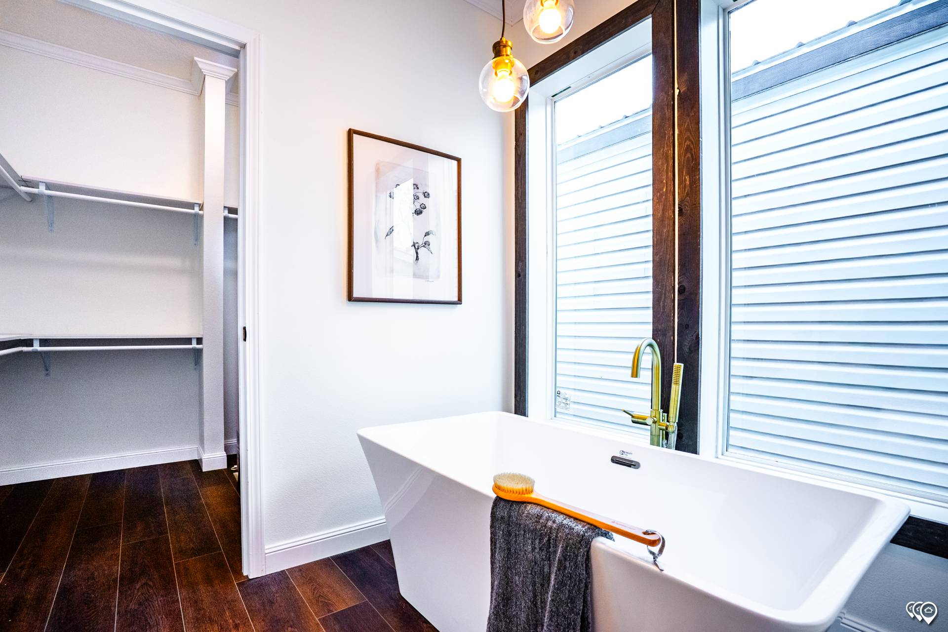 Minimalist bathroom with a white freestanding tub, gold faucet, and dark wood floor. A framed art piece and open closet add a modern touch.