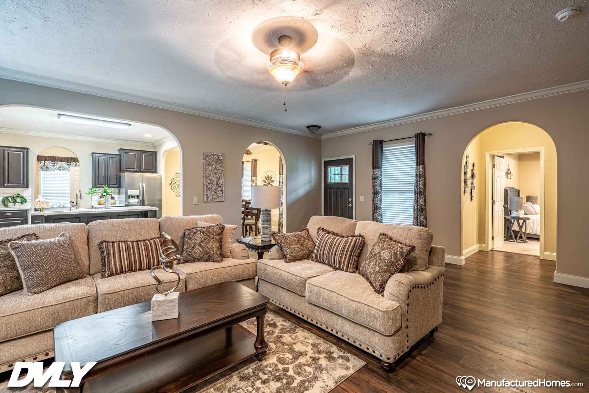 Spacious living room with beige sofas, dark wood coffee table, and cushions. Arched doorways lead to a kitchen and dining area. Ceiling fan and soft lighting create a warm, inviting atmosphere.