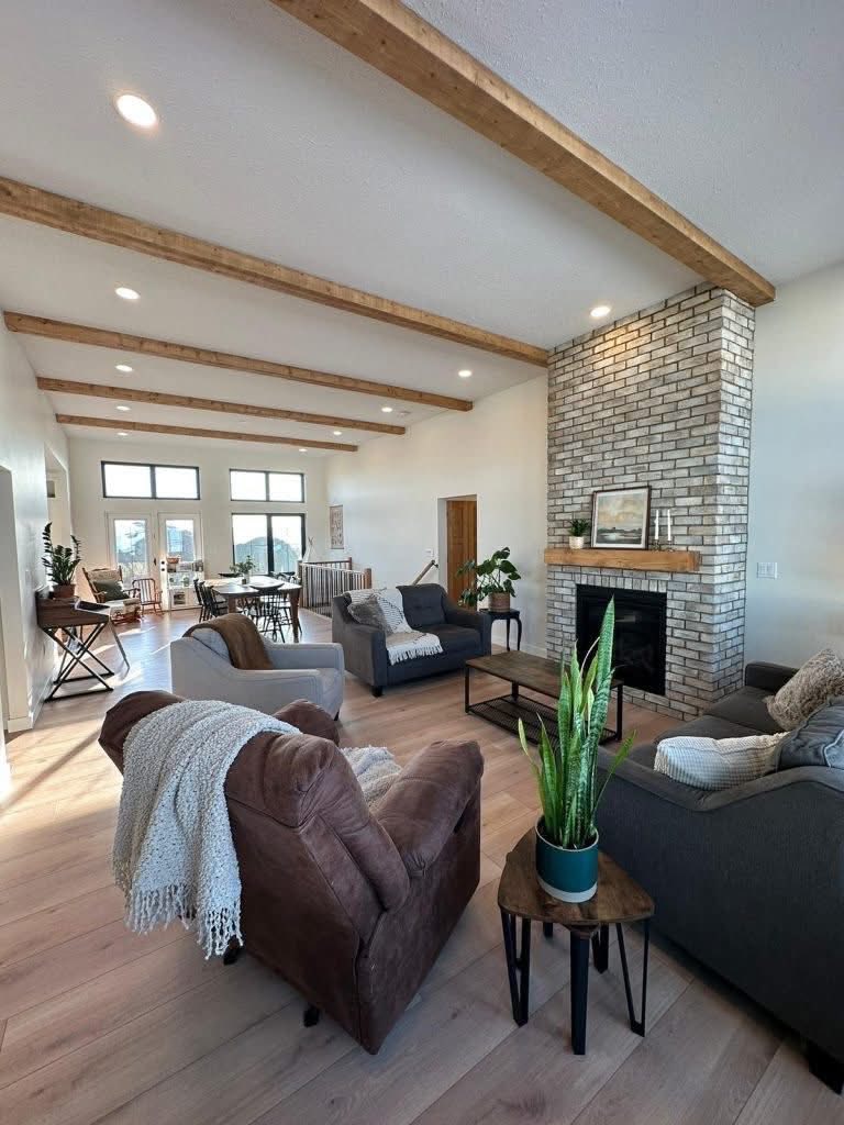Spacious living room with wooden beams, a brick fireplace, and cozy seating. Sunlit with plants, wooden floors, and a relaxed, inviting atmosphere.