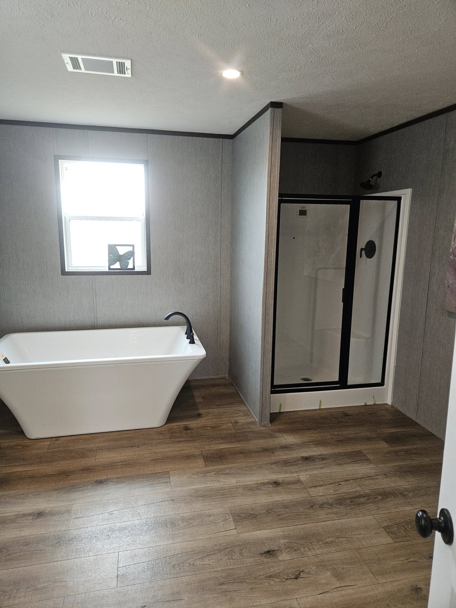Modern bathroom with a freestanding white tub, black faucet, and enclosed shower. Natural light streams through a window, highlighting wood floor.