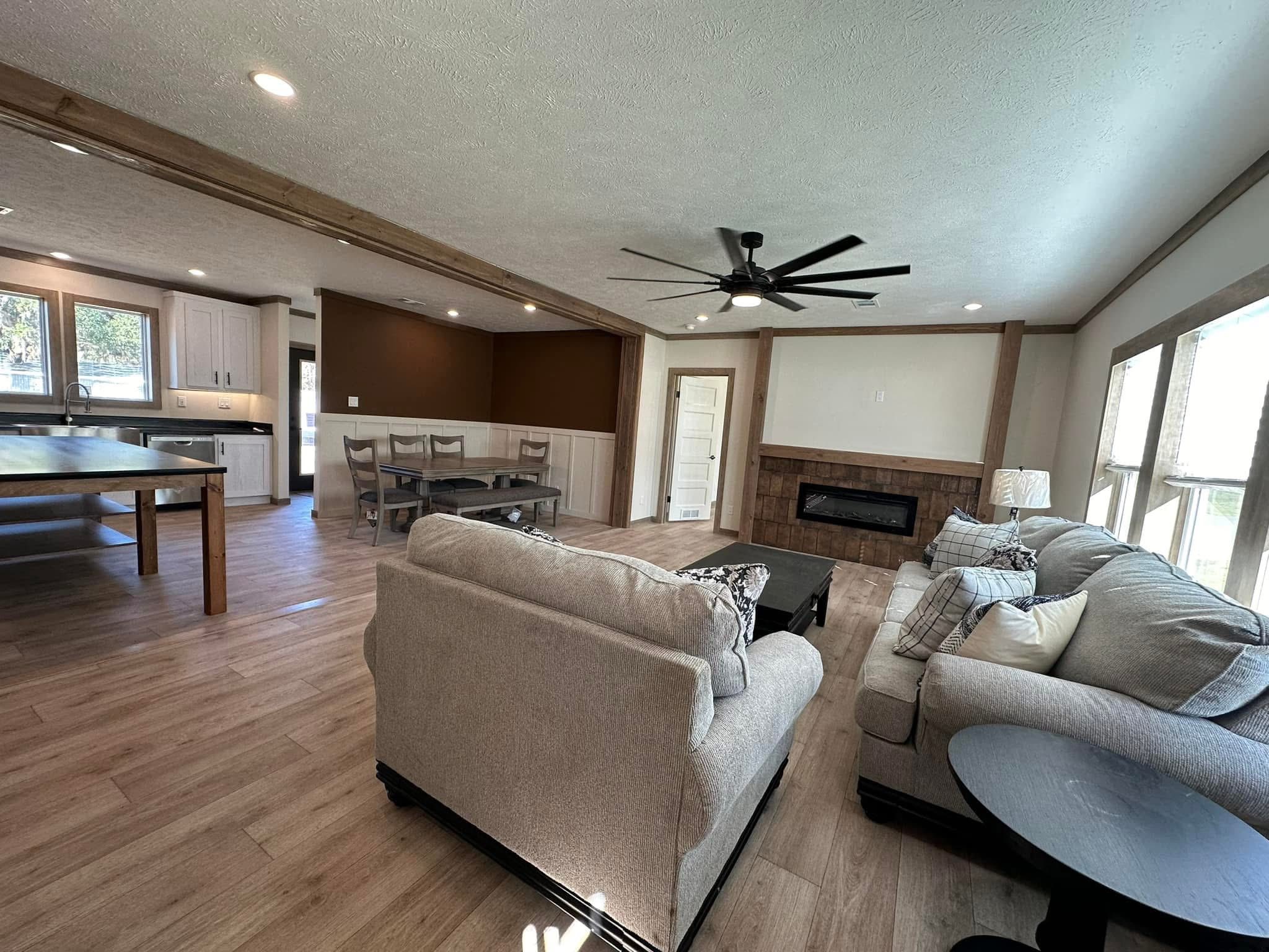 Modern open-plan living area with light wood flooring, neutral-toned sofas, dark ceiling fan, and large windows. Features a dining table and kitchen in the background.