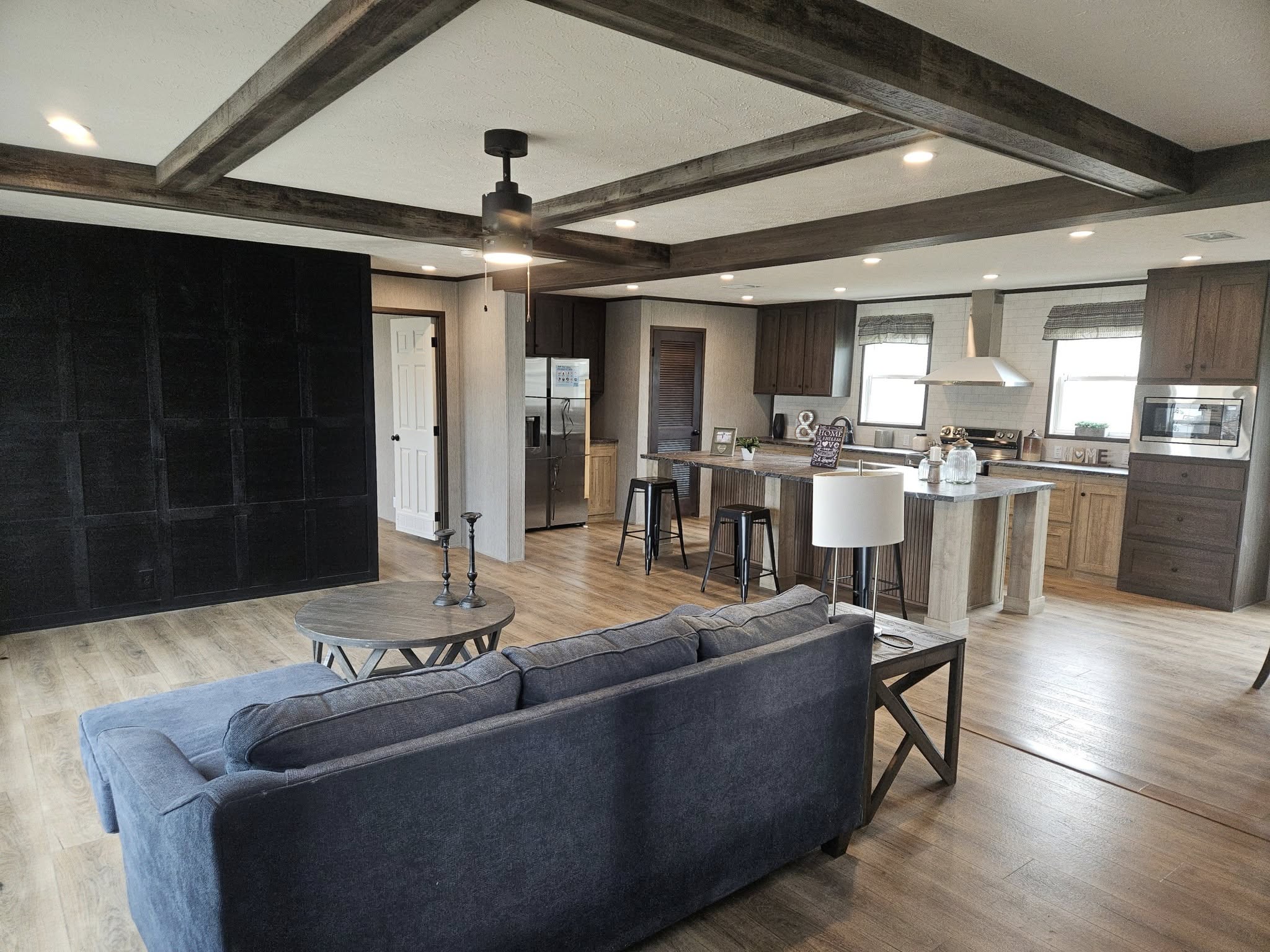 Spacious open-plan living room and kitchen. Features a dark blue sofa, wooden ceiling beams, modern lighting, a kitchen island with stools, and light wood floors. Cozy and contemporary atmosphere.