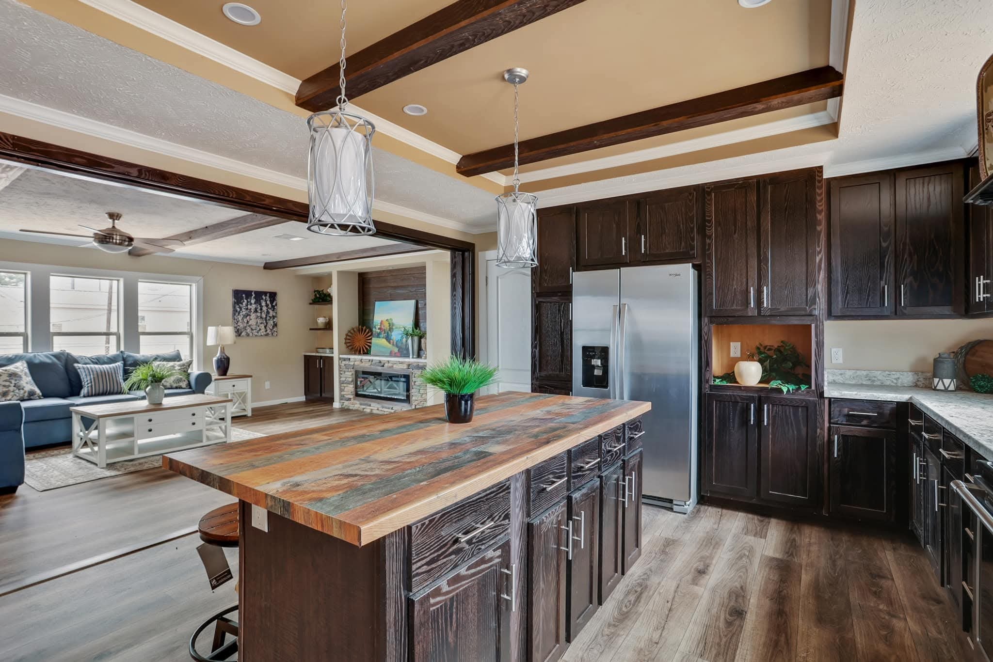 Modern kitchen with dark wood cabinetry and a rustic island. Stainless steel appliances, pendant lights, and a cozy living area in the background.