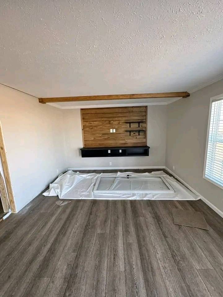 Minimalist room under renovation with wood floors, a wooden accent wall, and suspended black console. Plastic sheets and a window frame on the floor.