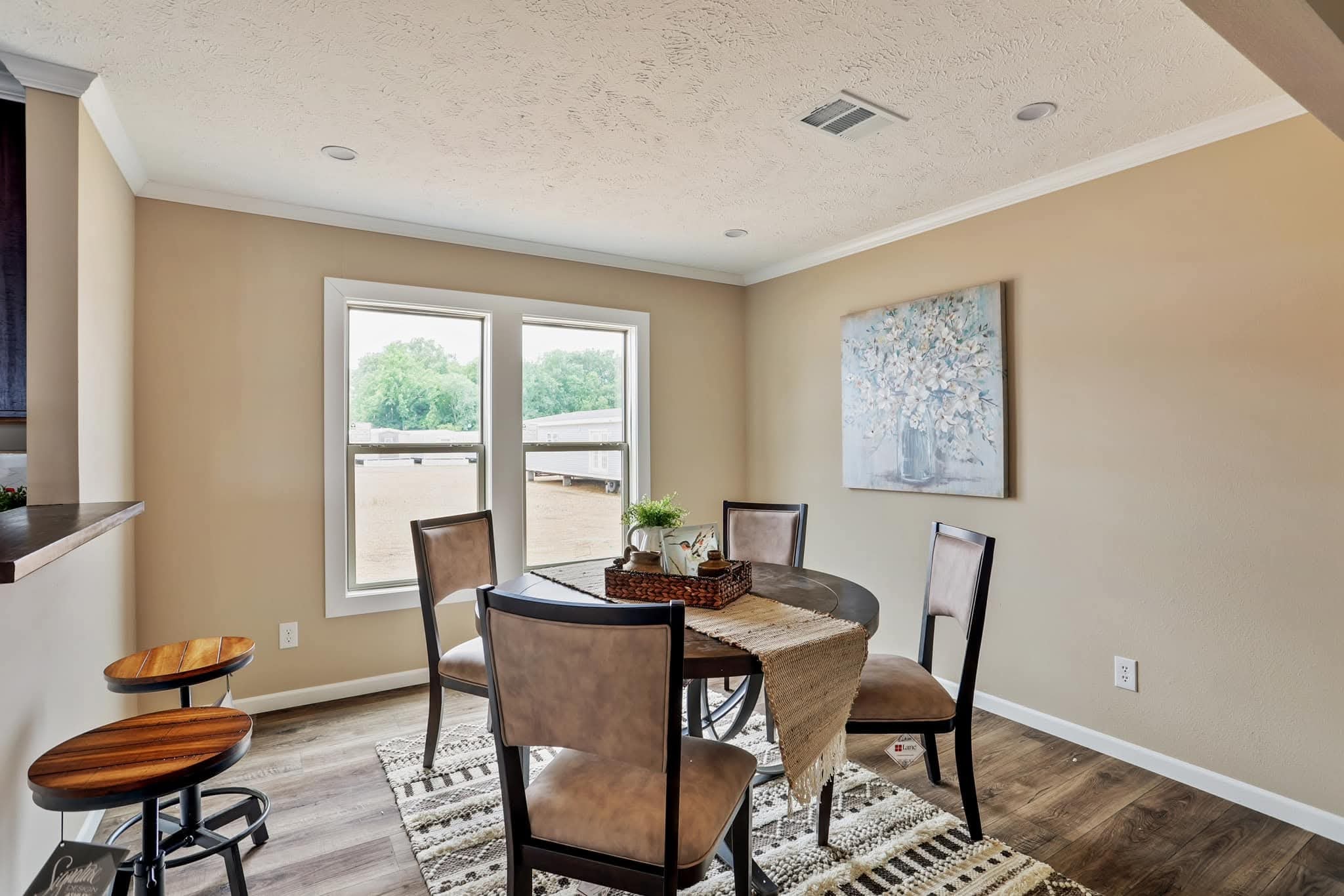 A cozy dining area featuring a round table with four cushioned chairs, a brown woven centerpiece, and a beige wall adorned with a floral painting. Natural light streams in through two large windows, highlighting the wood flooring and creating a warm, inviting atmosphere.