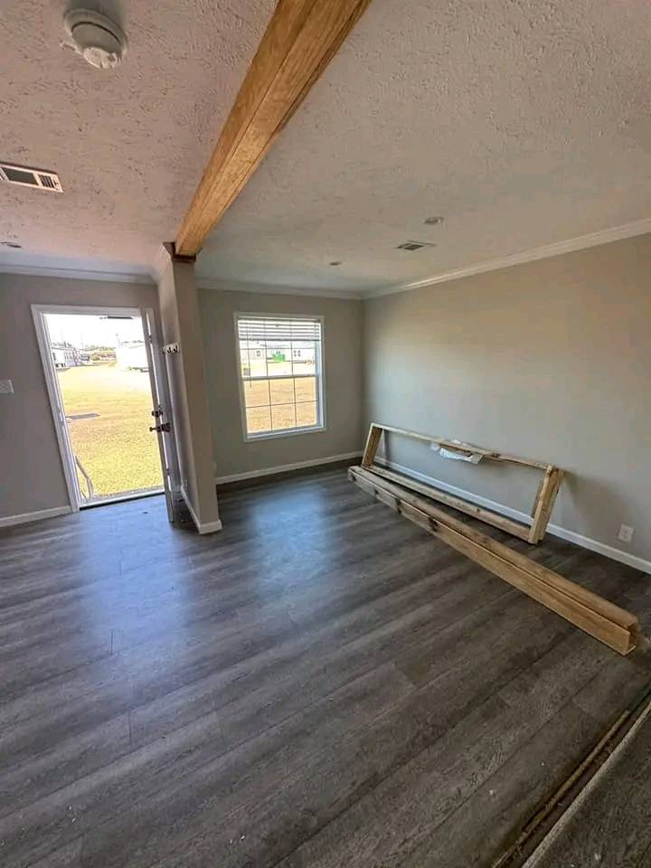 Empty room with light gray walls and wood-like flooring. A large window and open door let in natural light. Wooden beams are stacked against a wall.