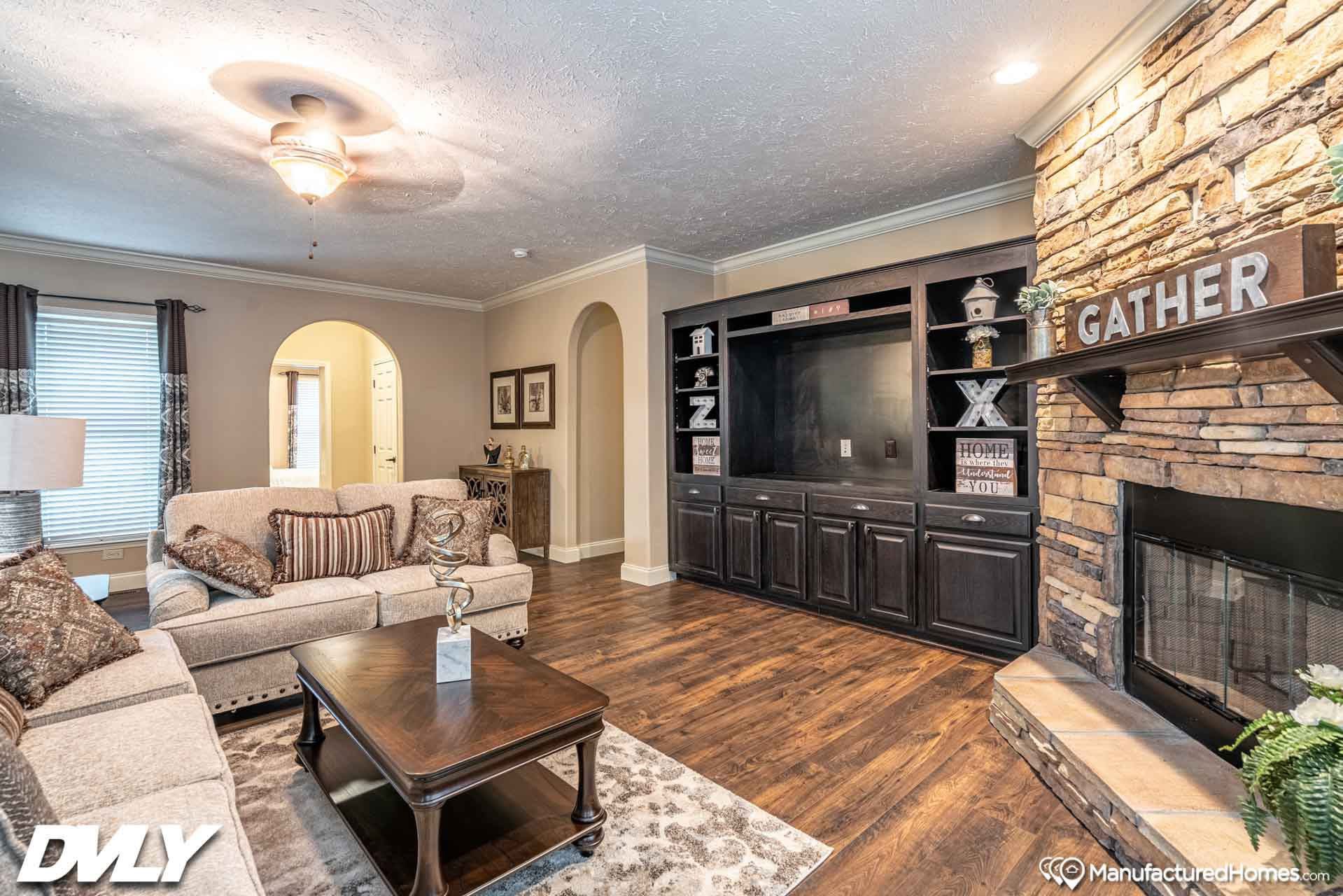 Warm and inviting living room with beige sofas, patterned cushions, and a wooden coffee table. A stone fireplace and dark wood shelves add coziness.