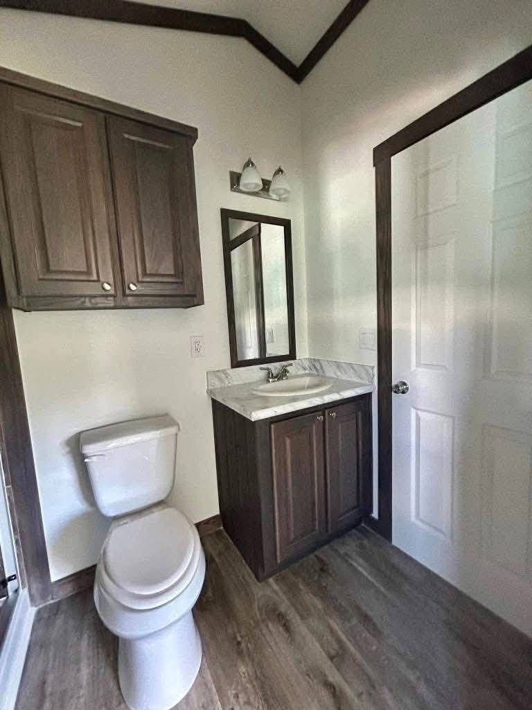 Small, modern bathroom with a toilet, wooden cabinet sink, wall mirror, and overhead light. White walls and wood floor create a clean, cozy tone.