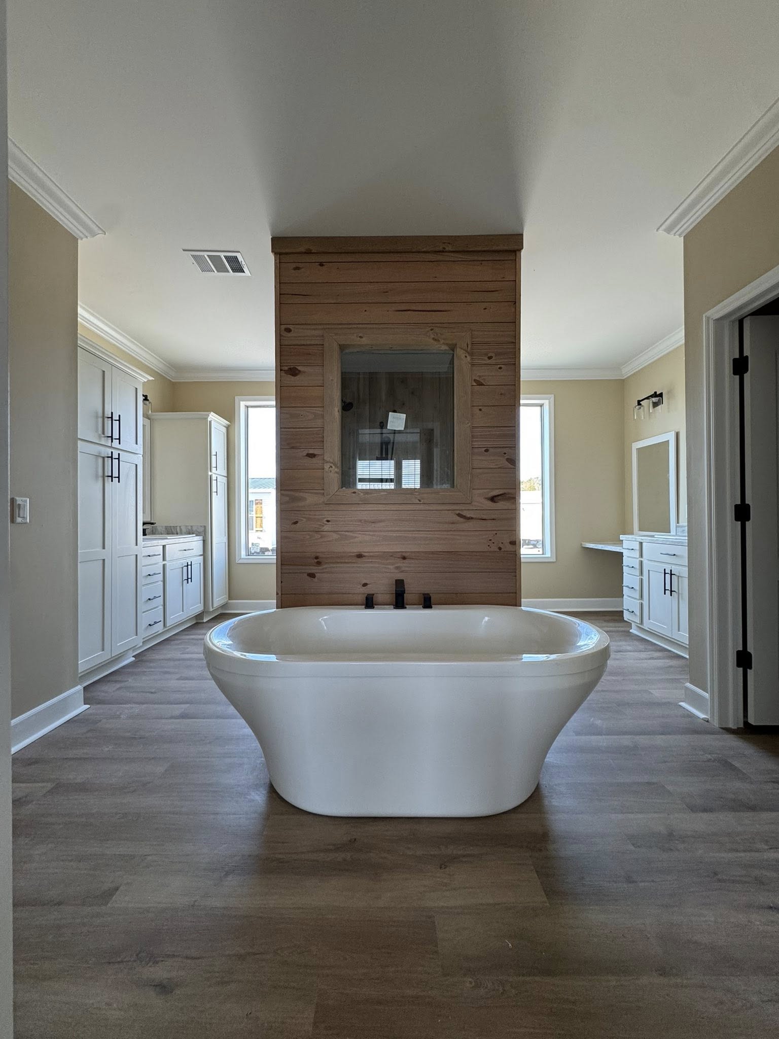 Freestanding white bathtub in center of a modern bathroom with wood paneling, two large windows, and neutral cabinetry. Bright, airy, and serene.