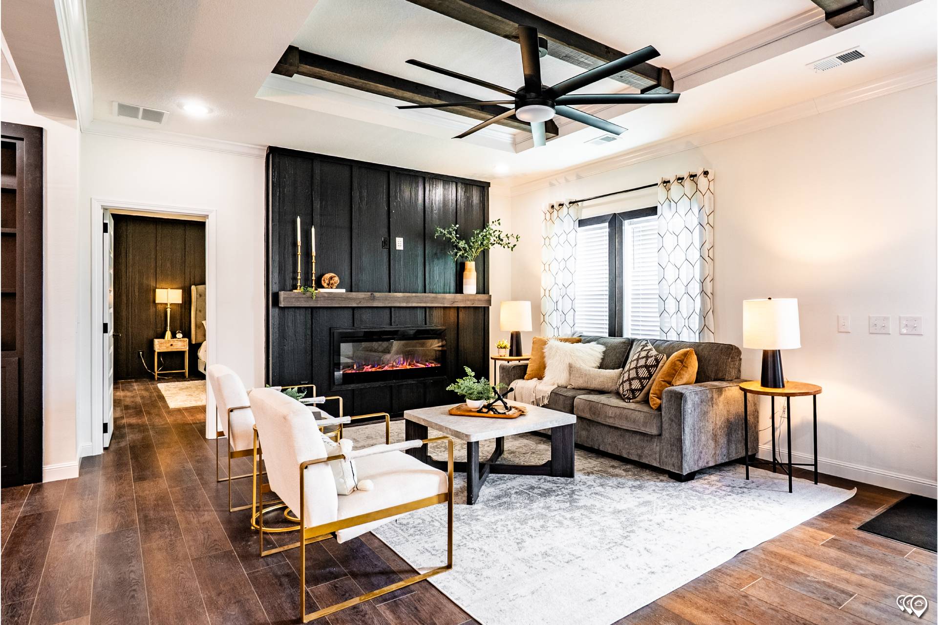 Stylish living room with wood floors, a dark wood fireplace, and gray sofa. Gold-accented chairs and a coffee table add elegance under a large ceiling fan.