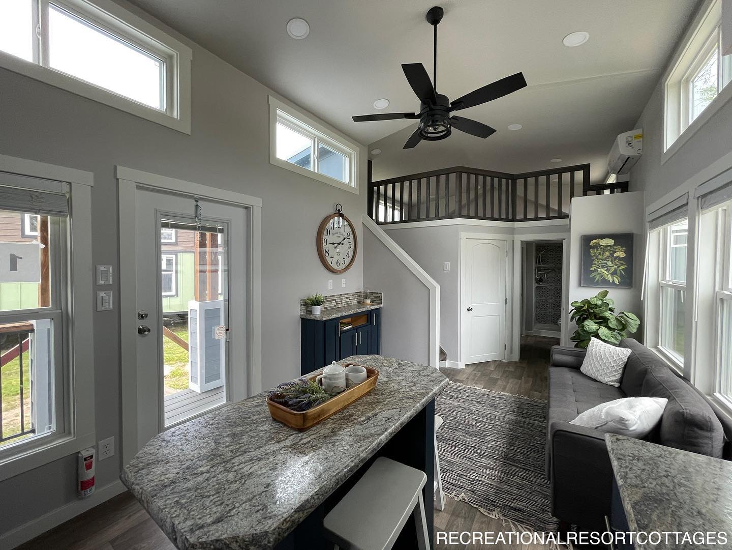 Modern tiny home interior with high ceilings, large windows, and a loft. Features a gray sofa, kitchen island with granite top, and decorative plants. Cozy and bright ambiance.