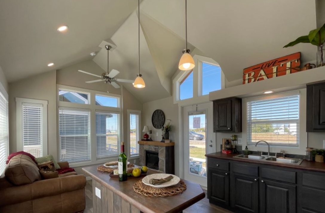 Cozy kitchen and living area with vaulted ceilings, pendant lights, and large windows. Features a brown sofa, island with fruit, and dark wood cabinets.