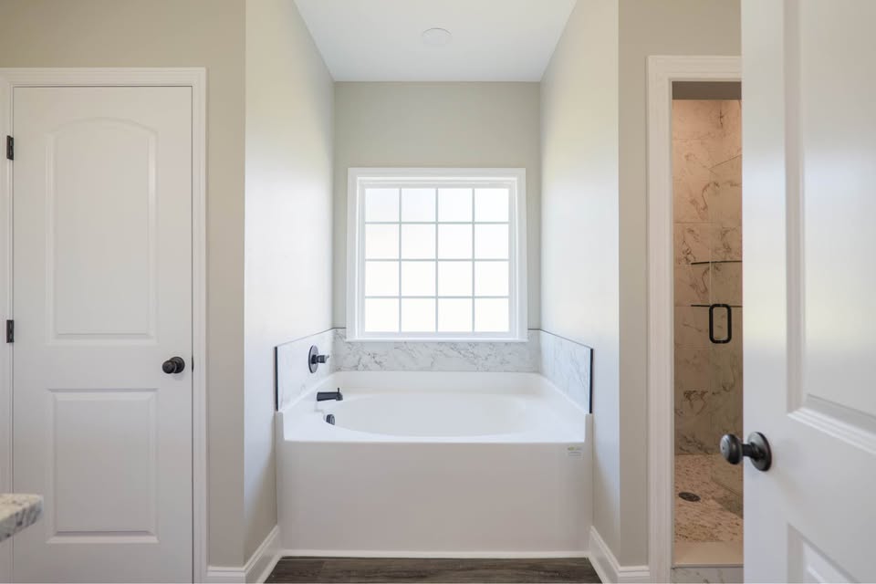 Bathroom with a white bathtub under a window, between beige walls. Adjacent are two doors, one leading to a marble-tiled shower space. Modern and serene.