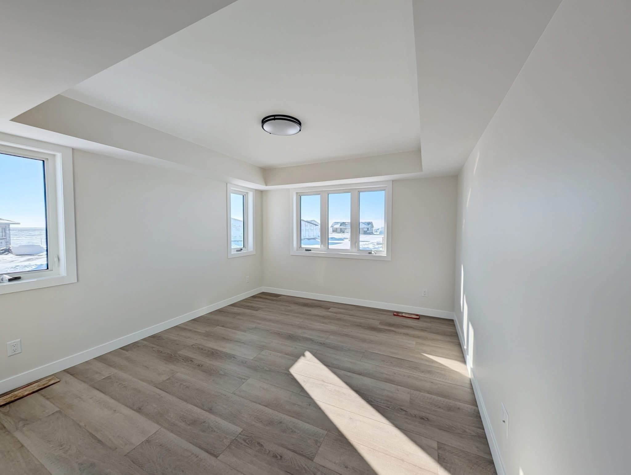 An empty, sunlit room with light wood floors and white walls featuring a modern ceiling light. Three large windows offer a view of snowy rooftops.
