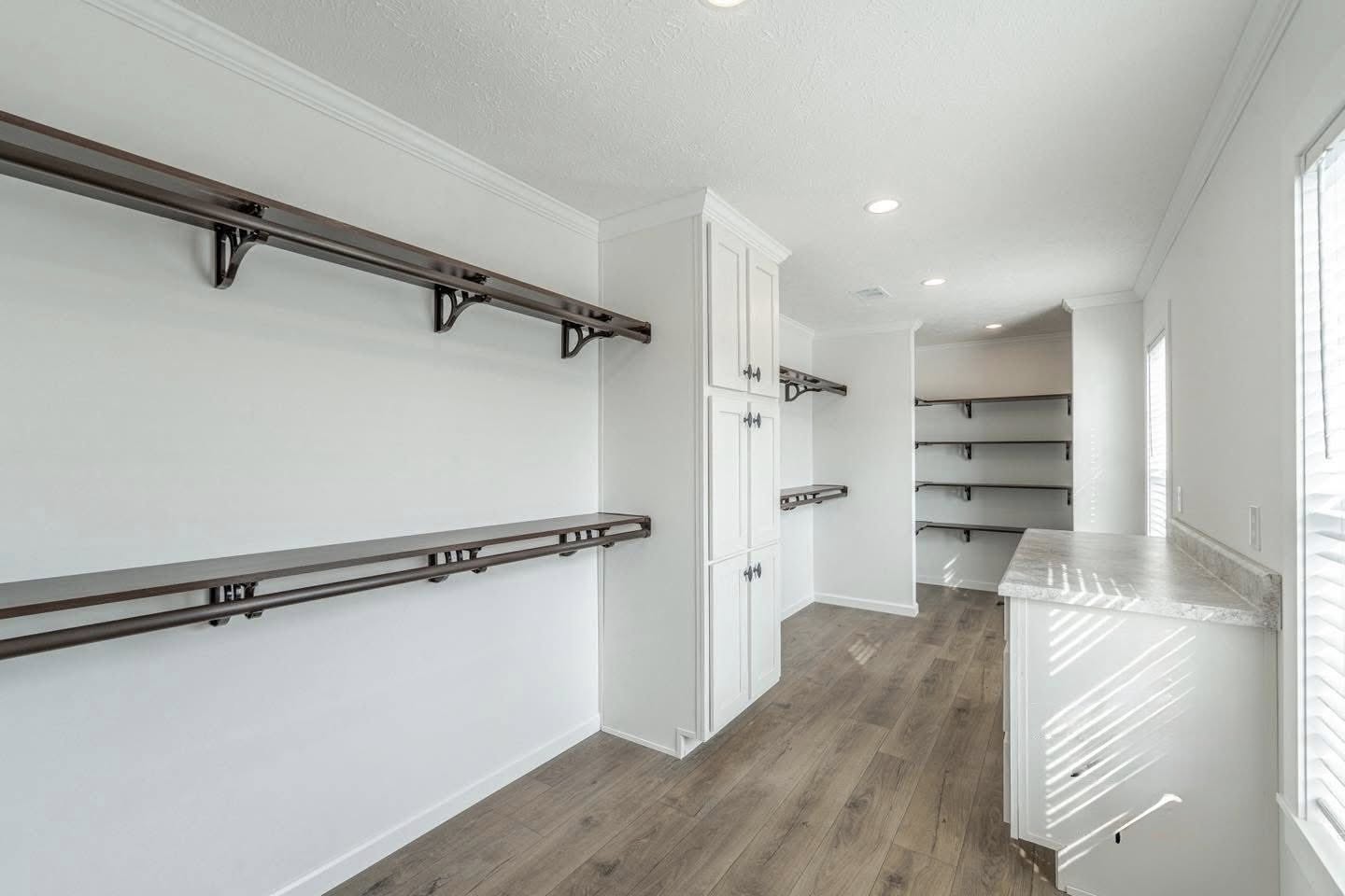 Spacious walk-in closet with white walls, wooden shelves, and cabinets. Natural light streams through a window. Clean and organized atmosphere.