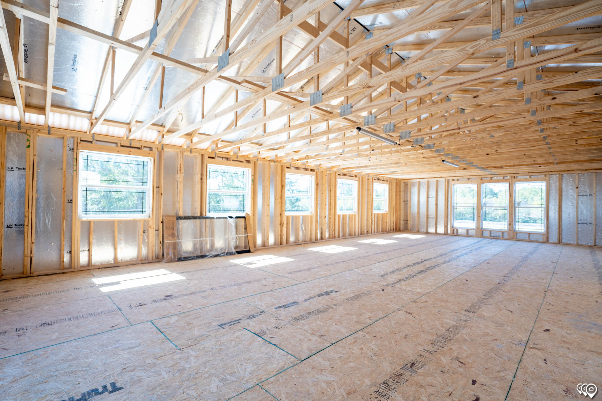 A sunlit, unfinished room with wooden framing, open truss ceiling, and large windows. The space feels spacious and airy, suggesting new construction.