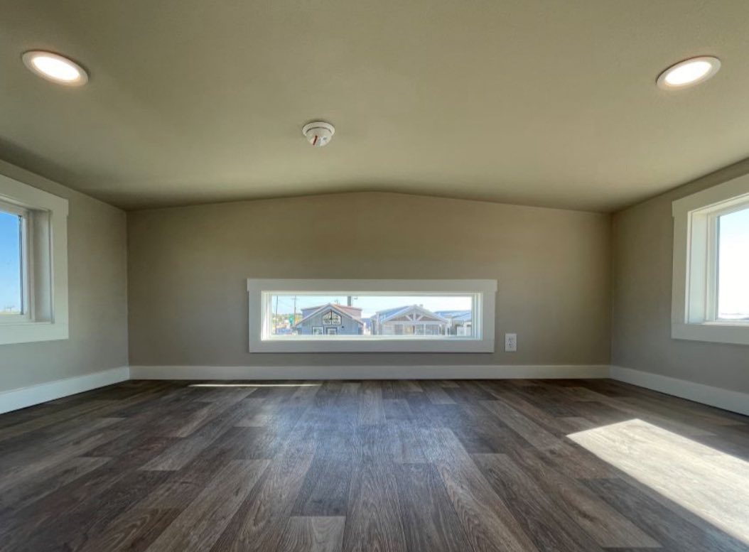 A small, empty attic room with dark wooden flooring and beige walls. Three rectangular windows let in bright sunlight, creating a cozy, quiet ambiance.