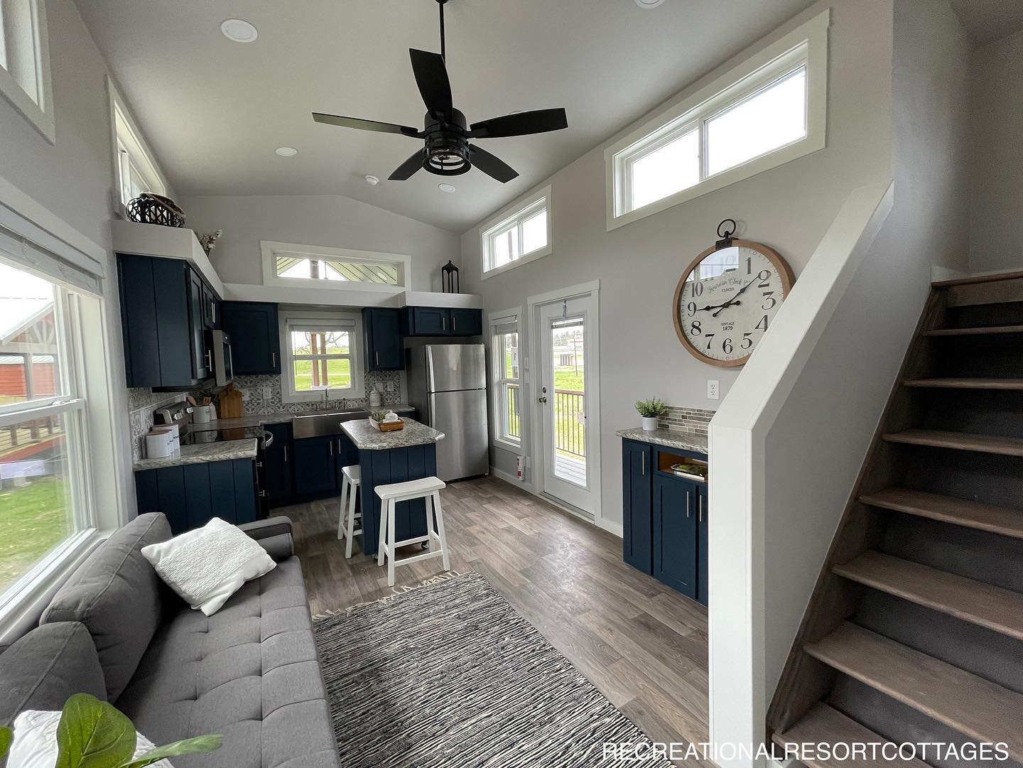 A modern tiny house interior with a cozy living area, gray sofa, and textured rug. The compact kitchen has blue cabinets and a small island. Large windows bring in natural light. An eye-catching clock decorates the wall near the staircase, and a ceiling fan hangs above, creating an inviting and spacious feel.