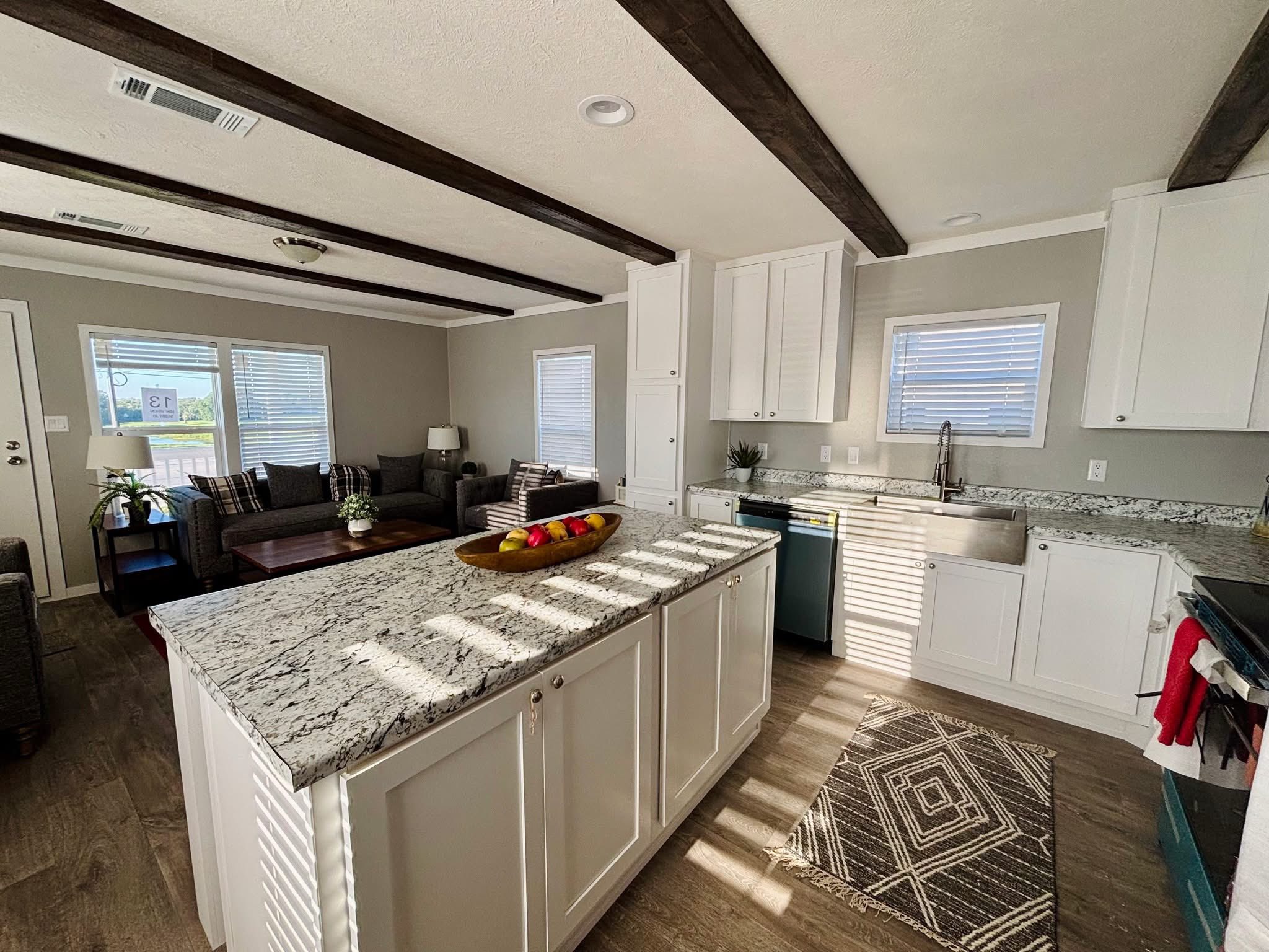 Modern kitchen with a marble island and white cabinets, leading to a cozy living area with gray sofas. Sunlight streams through multiple windows, creating a warm and inviting atmosphere.