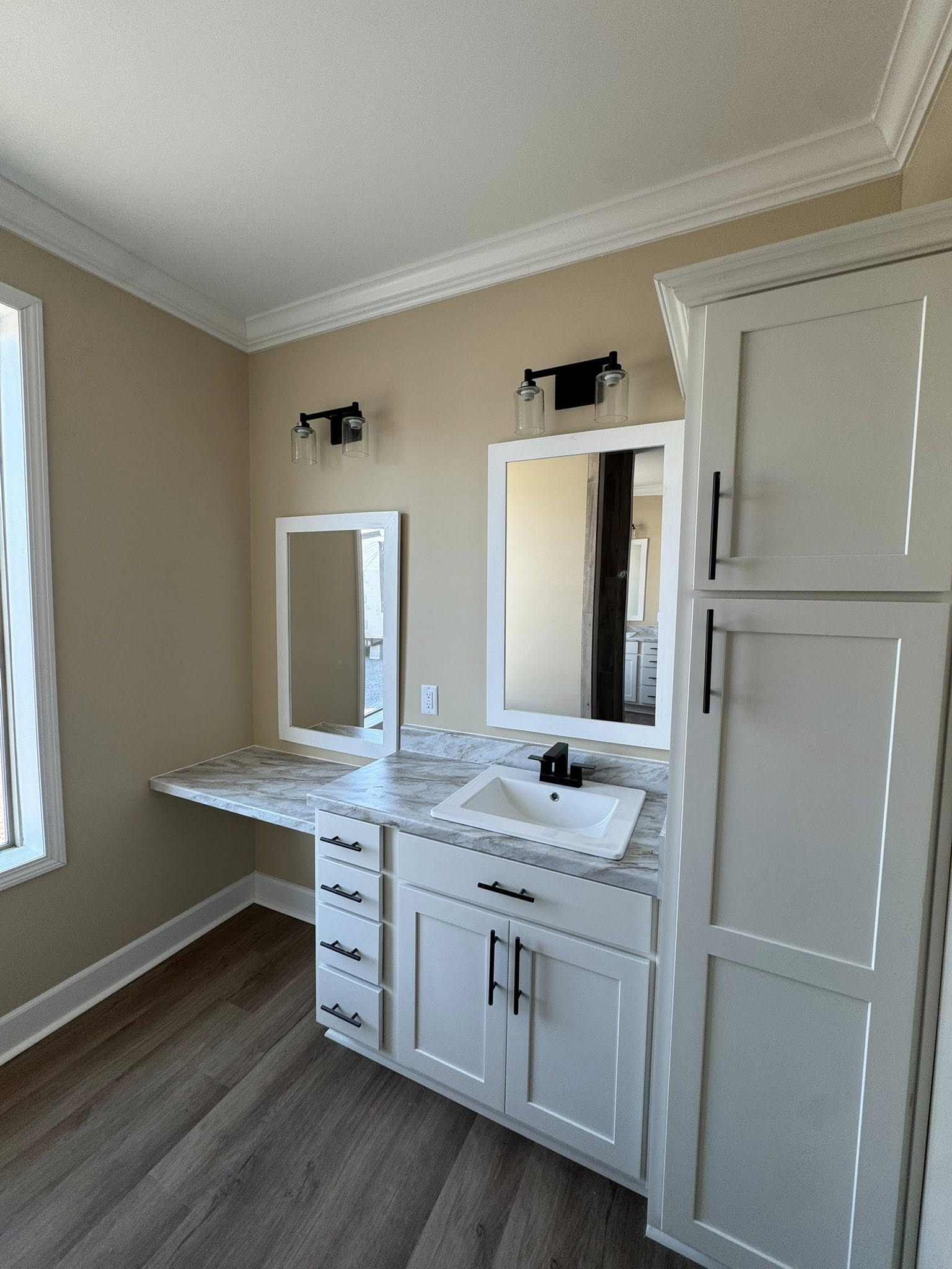 Modern bathroom with beige walls, white cabinets, and gray-veined countertop. A rectangular sink and two mirrors are visible. Natural light streams in.