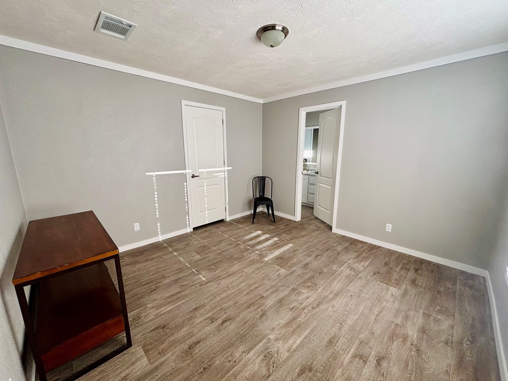 Sparse room with gray walls, wooden floor, and two white doors. A black chair sits near one door, and a dark wooden table is against the opposite wall.