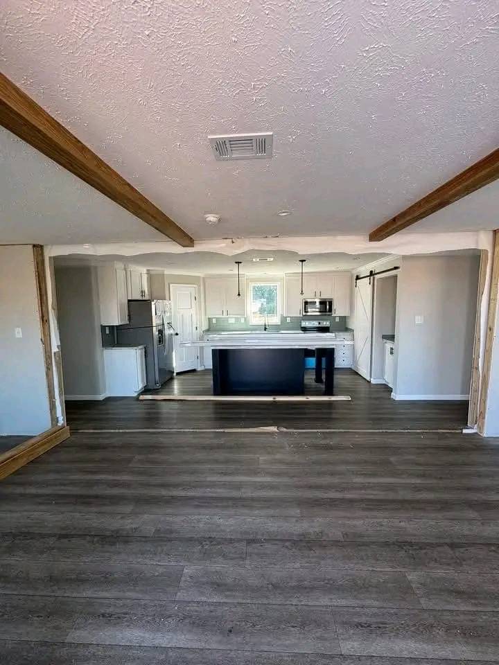 Spacious open-plan kitchen with a central island, dark wood floors, white cabinets, stainless steel appliances, and exposed wooden ceiling beams.
