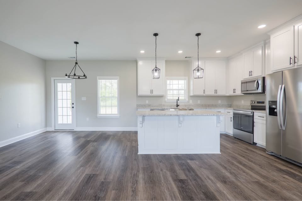 Modern kitchen with white cabinets, stainless steel appliances, and a central island under pendant lights. Wood flooring adds warmth. Bright, airy feel.