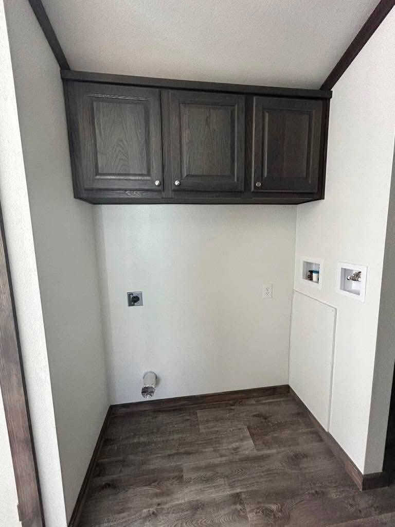 Empty laundry nook with dark wood cabinets above, white walls, and brown wood flooring. Plumbing hookups are visible, conveying a clean, unfinished look.