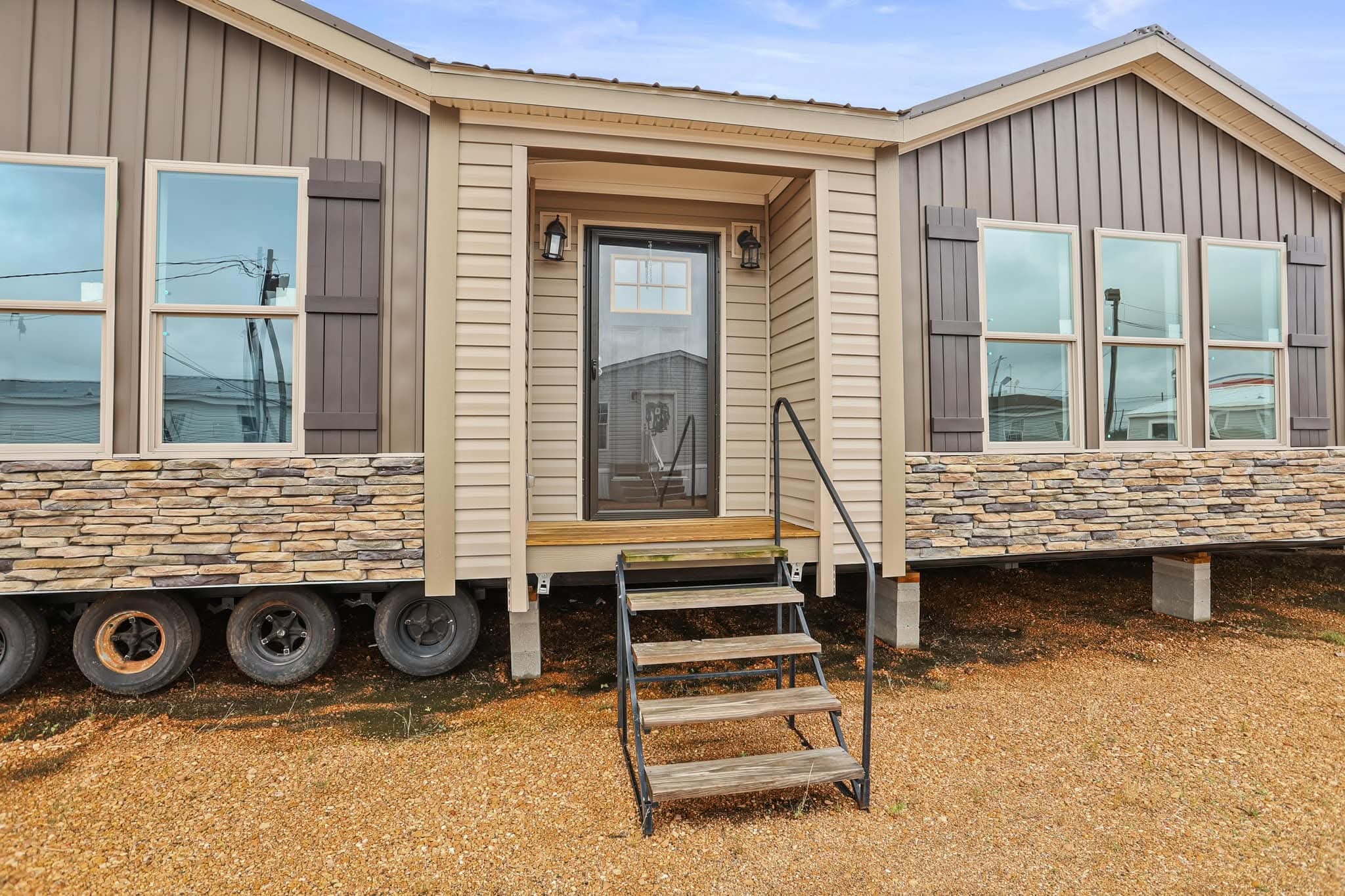 A modern mobile home with stone paneling and beige siding features large windows. It is supported by wheels, with steps leading to the glass front door.