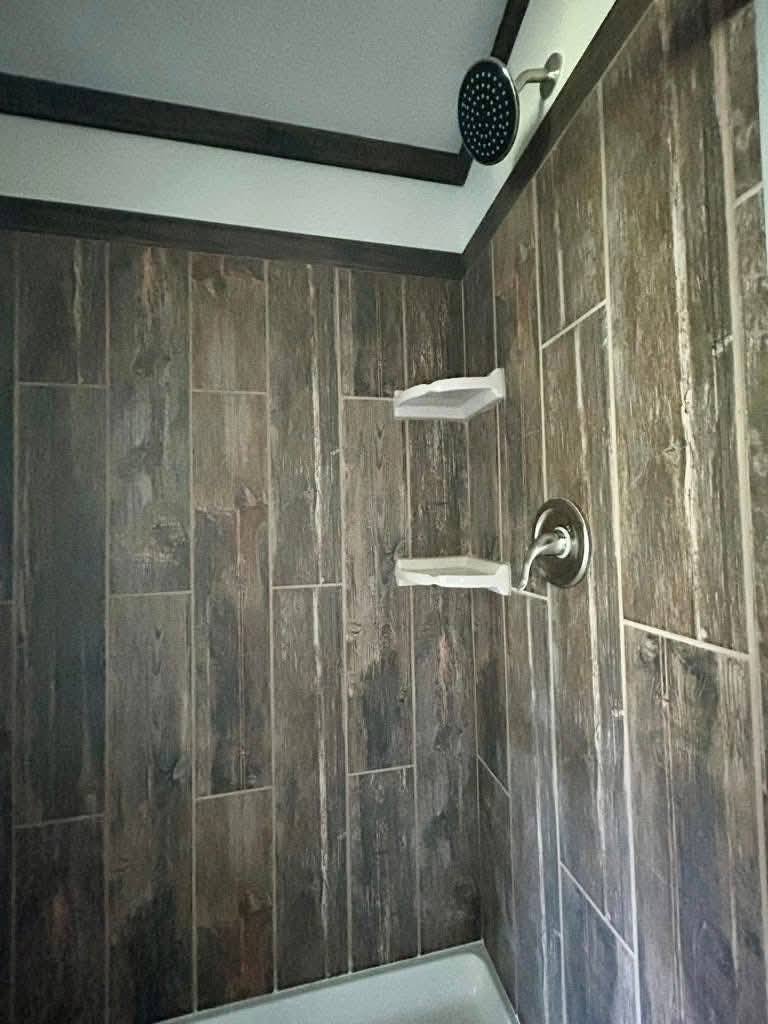 Shower with dark wood-patterned tiles, two white corner shelves, and a round showerhead. The atmosphere is modern and rustic.