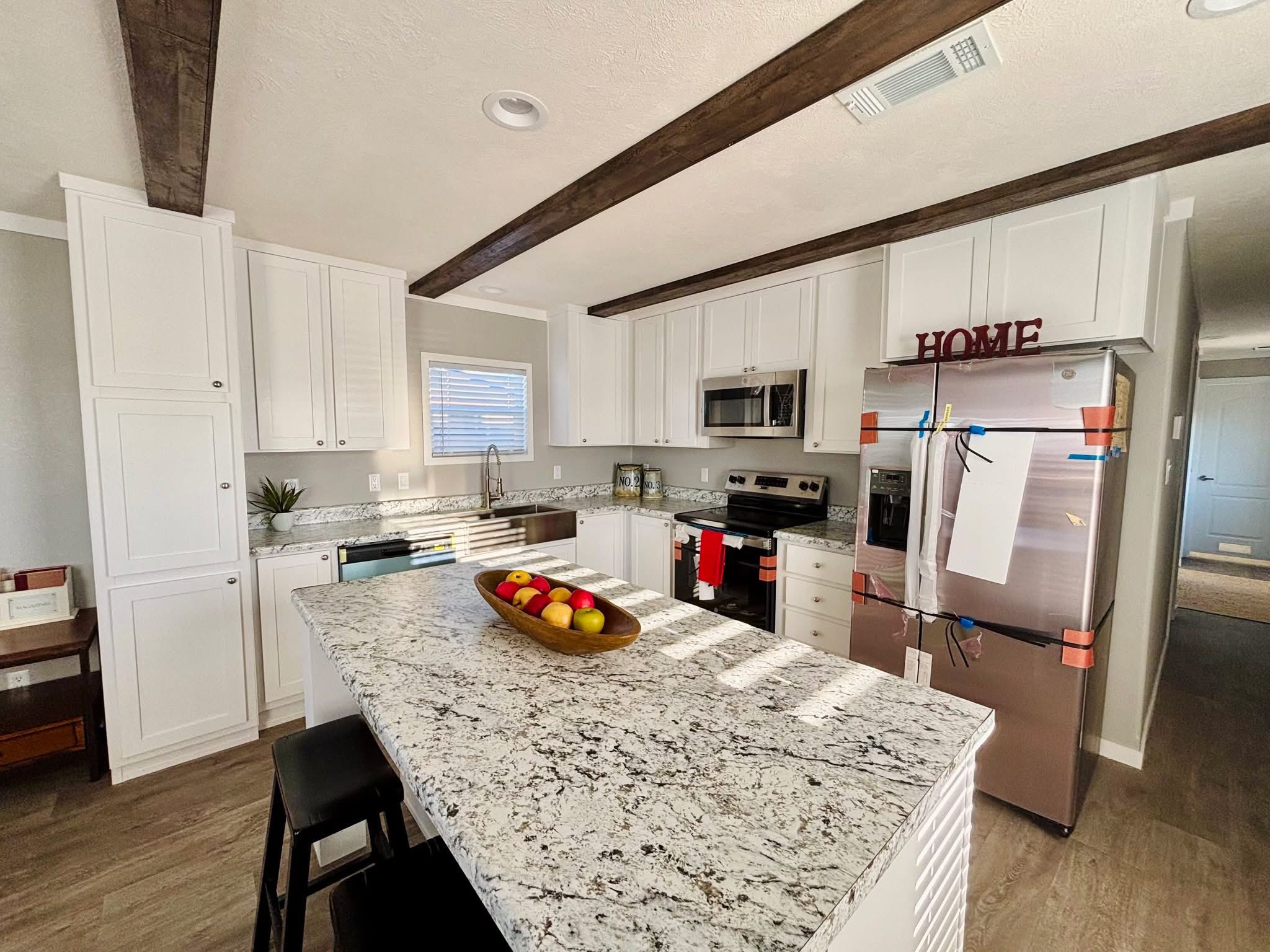 Modern kitchen with white cabinets, granite island, and stainless steel appliances. A bowl of colorful fruit sits on the island, and "HOME" decor atop the fridge adds warmth.