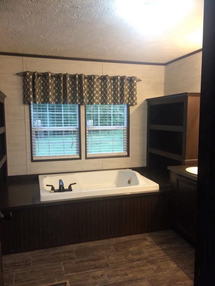 Cozy bathroom with a large white bathtub beneath two windows with blinds and patterned curtains. Dark wooden shelves flank one side, creating a warm ambiance.
