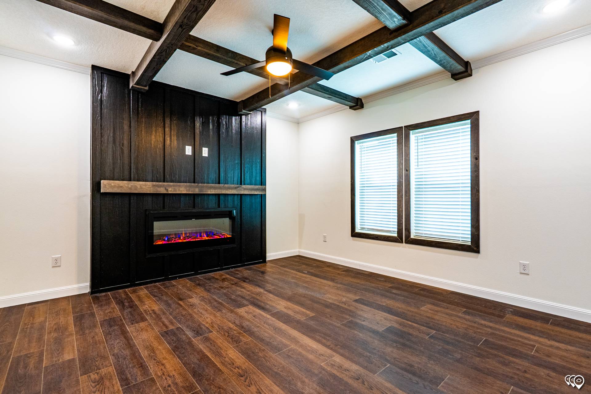 Spacious room featuring a dark wood accent wall with an electric fireplace, ceiling beams, fan, large windows, and warm-toned wooden flooring.