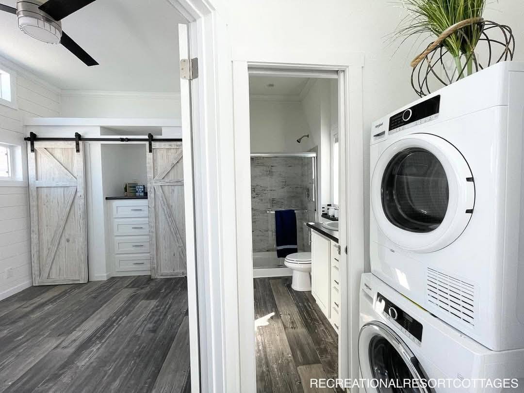 Modern laundry area with stacked washer and dryer beside a bathroom with a tiled shower and blue towel. Adjacent room features sliding barn doors.