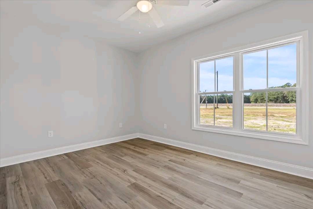 An empty room with light gray walls and wood flooring. A large window on the right shows a grassy field outside. A white ceiling fan is overhead.