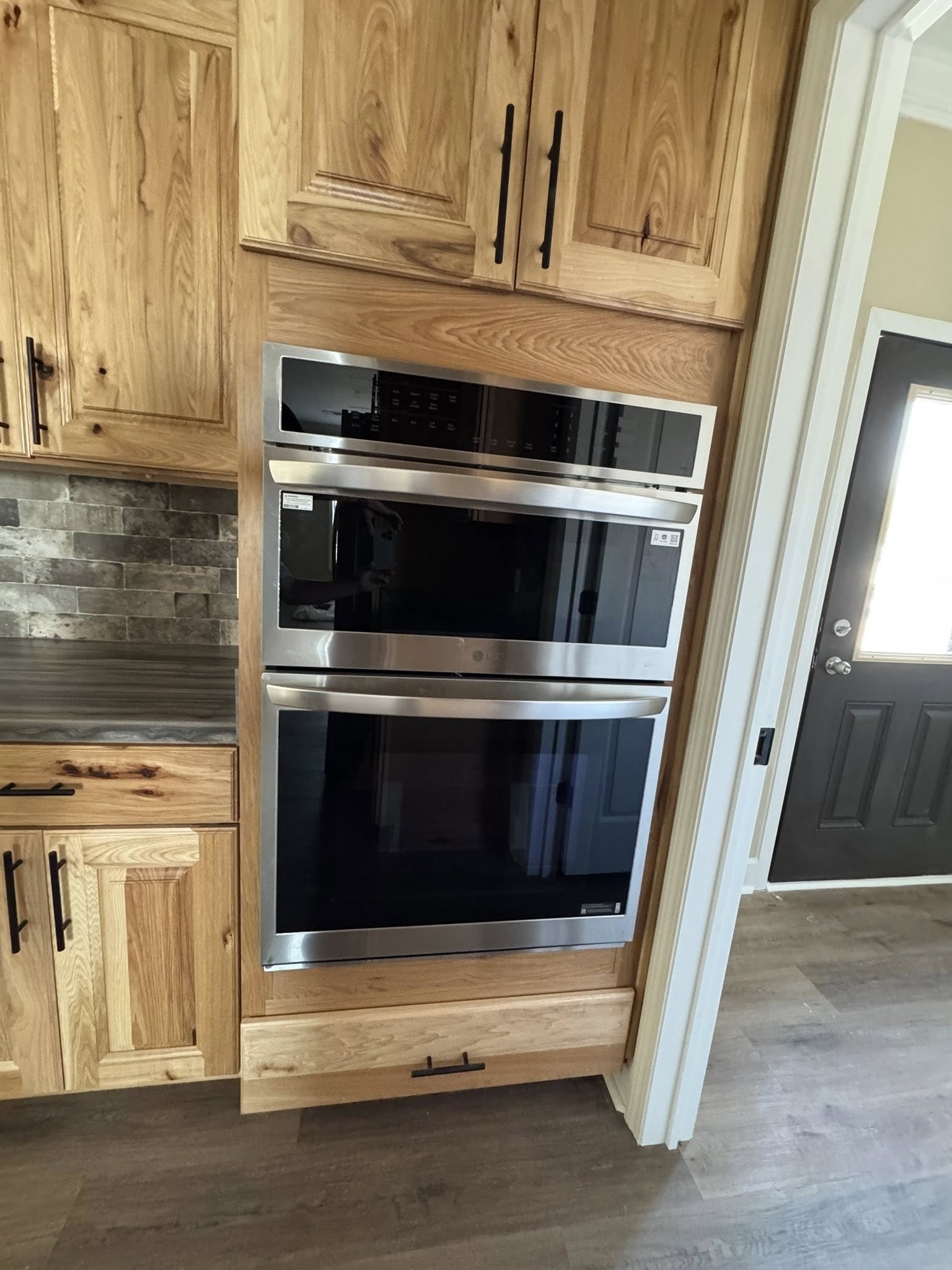 Wooden cabinetry with modern built-in double ovens and sleek metal handles. Beside them, a dark tile backsplash contrasts the light wood tones.