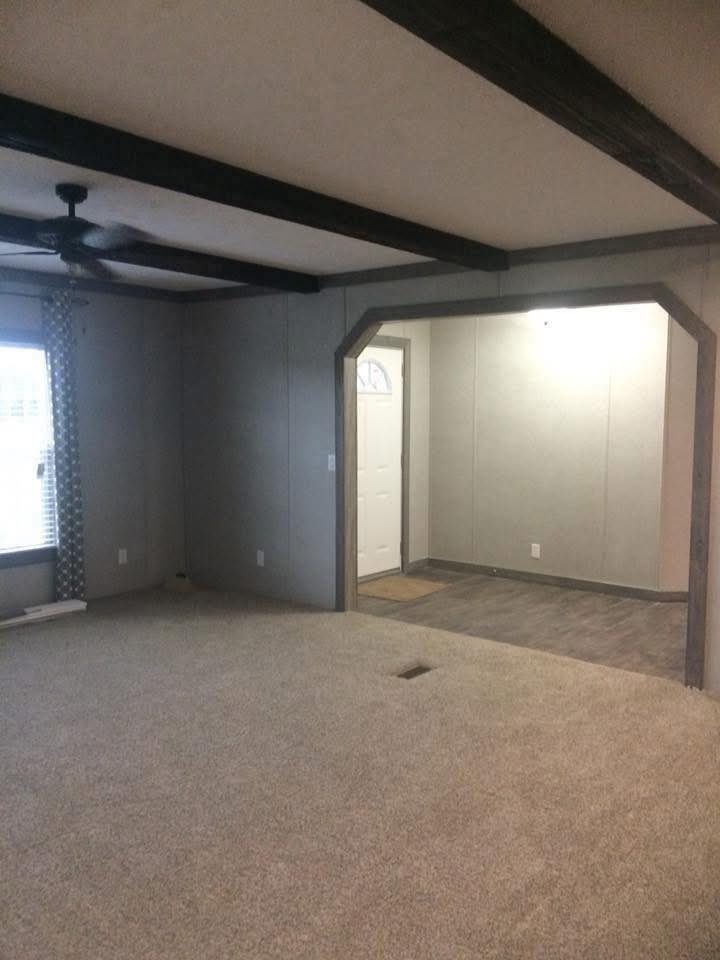 A room with gray walls and ceiling beams features a carpeted floor, a ceiling fan, and a window with patterned curtains. An entrance leads to a smaller area with wood flooring, under soft lighting, giving an inviting feel.