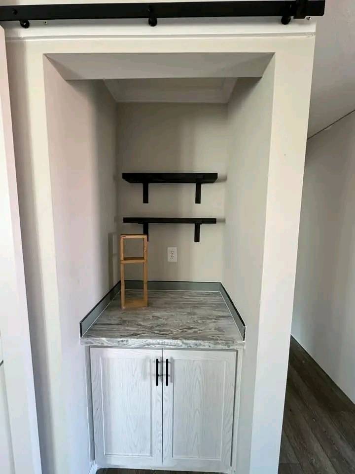 Small recessed nook with two black floating shelves. Below, a gray marble countertop and white cabinet with black handles. Wooden floor visible.