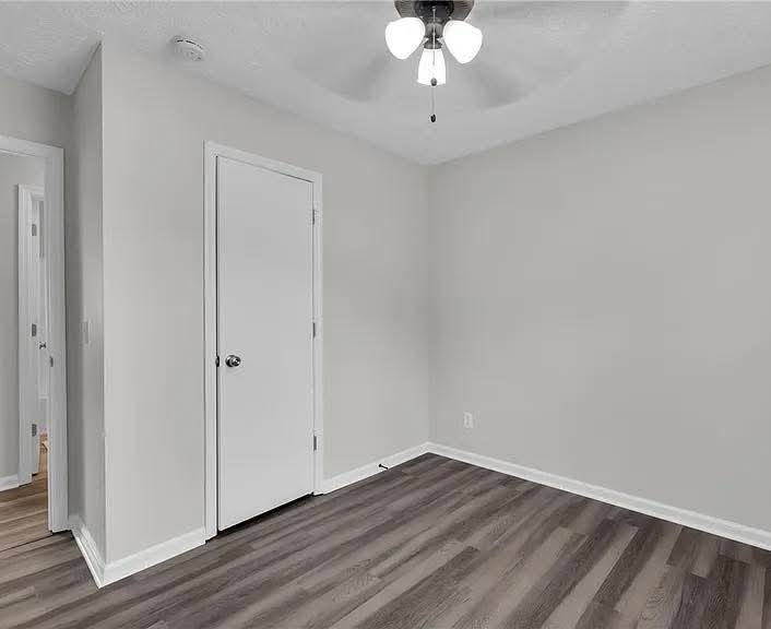 Empty room with light gray walls, a white door, and dark wood laminate flooring. A ceiling fan with lights hangs, creating a minimalist, clean atmosphere.