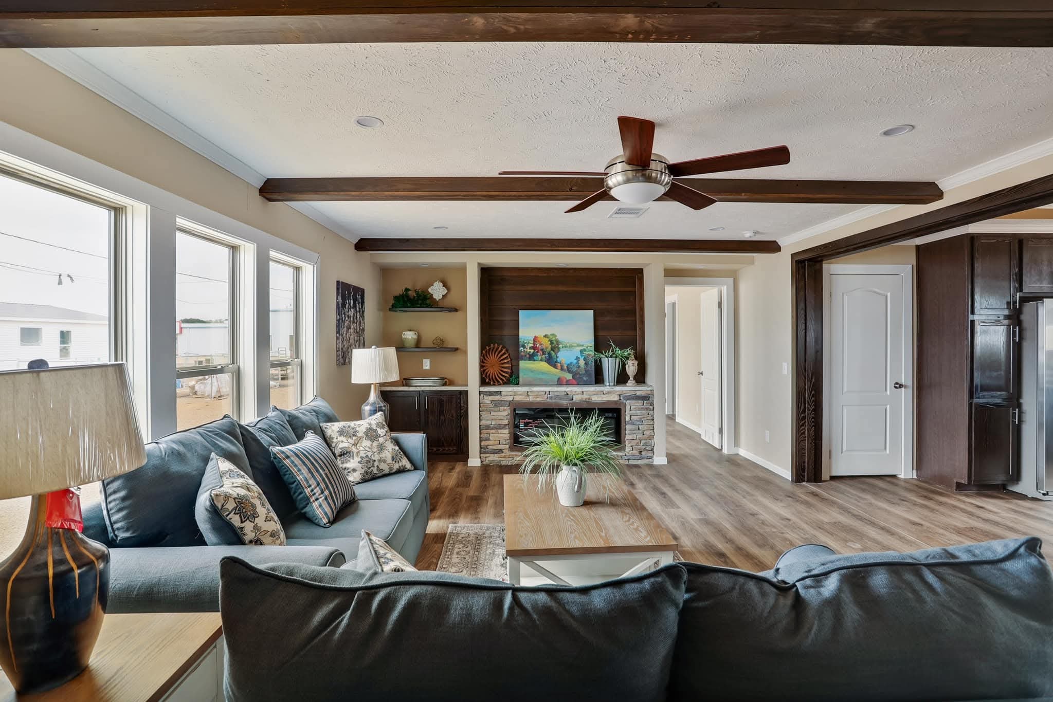 Cozy living room with light wood flooring, blue sofas, decorative pillows, ceiling fan, fireplace, artwork, and potted plants. Bright, inviting atmosphere.