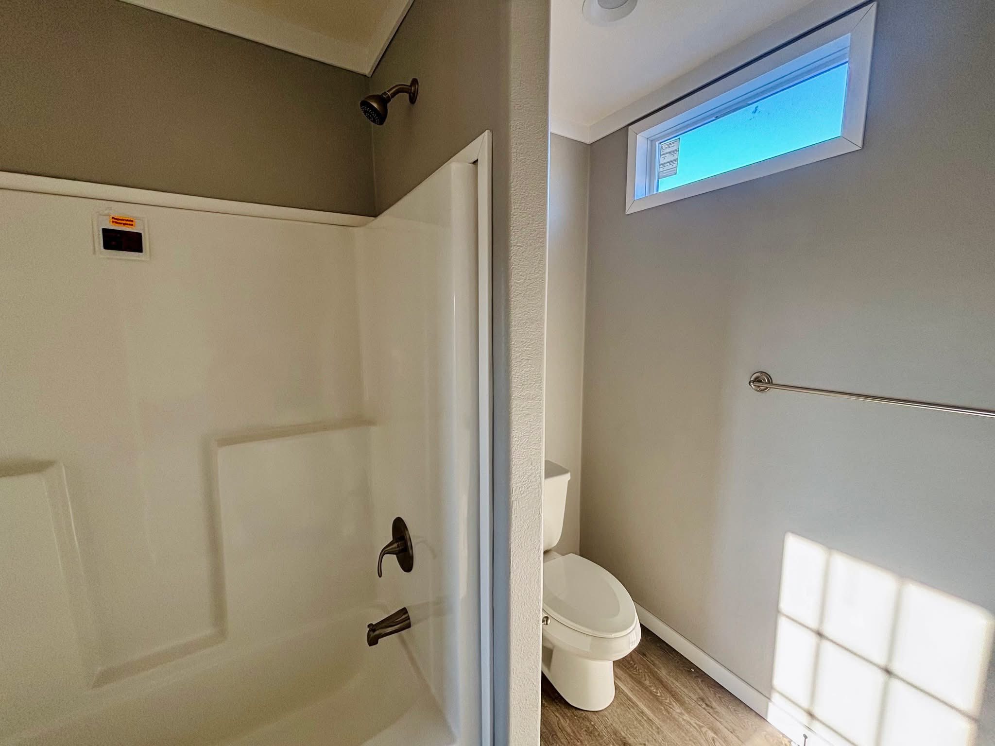 Compact bathroom with gray walls and wooden floor. Features a shower with bronze fixtures, a white toilet, and a small window allowing sunlight in.