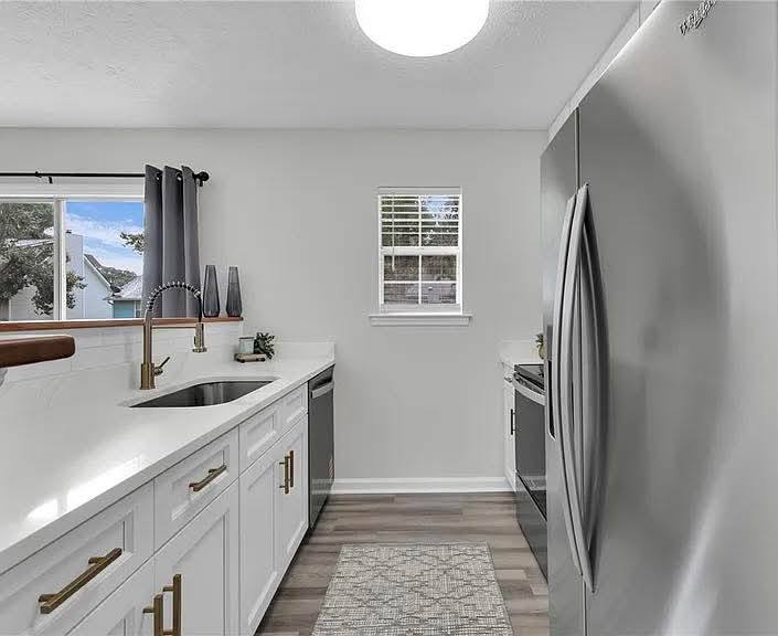 Modern kitchen with gray and white decor. Features stainless steel fridge, white cabinets with gold handles, and a window with gray curtains. Bright and clean.