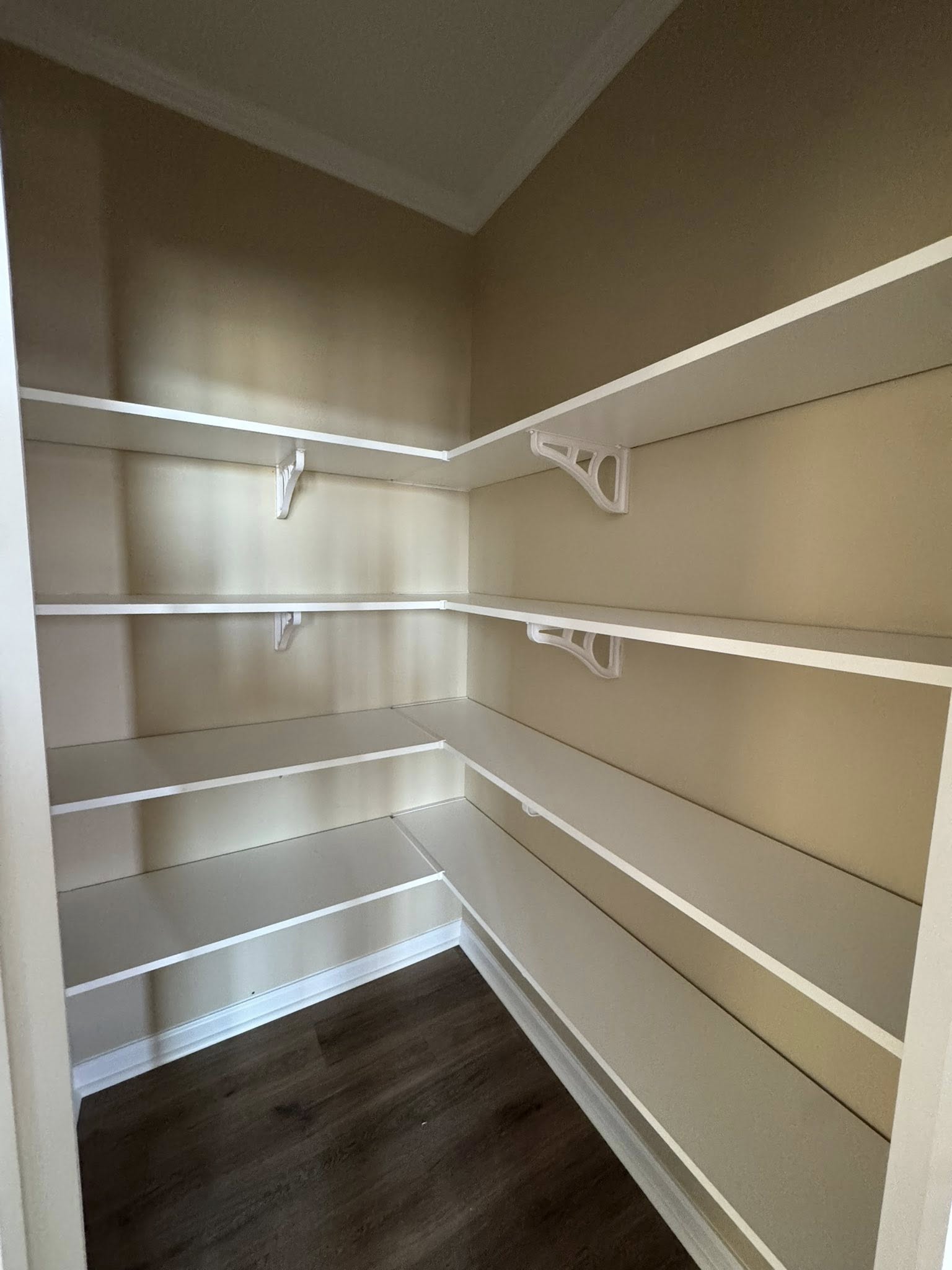 Empty pantry with beige walls, featuring five white shelves supported by brackets. The wooden floor and soft lighting create a clean, organized feel.