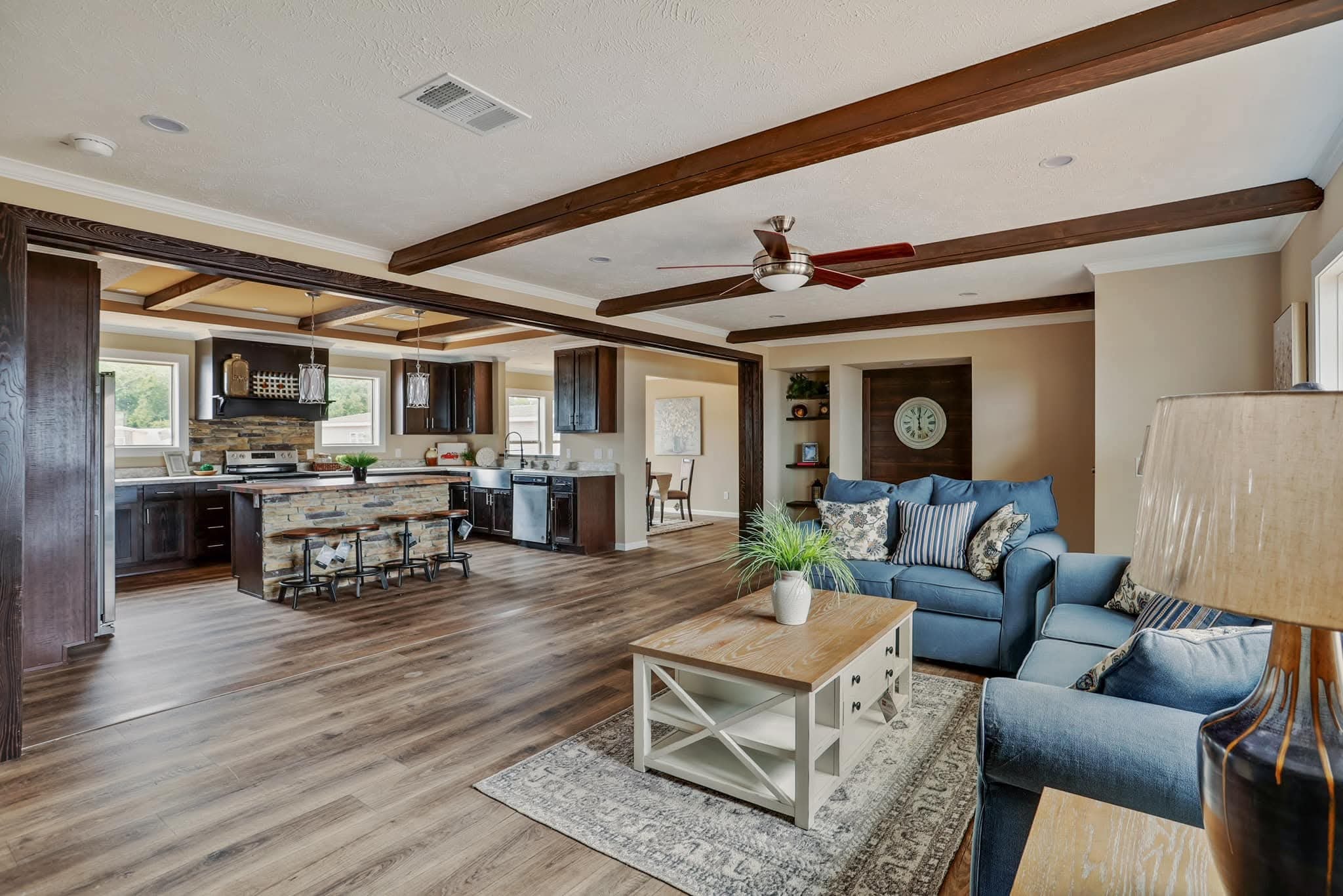 Open-concept living room and kitchen with wooden beams, blue sofas, and a rustic coffee table. The kitchen features an island with stools. Bright and cozy.