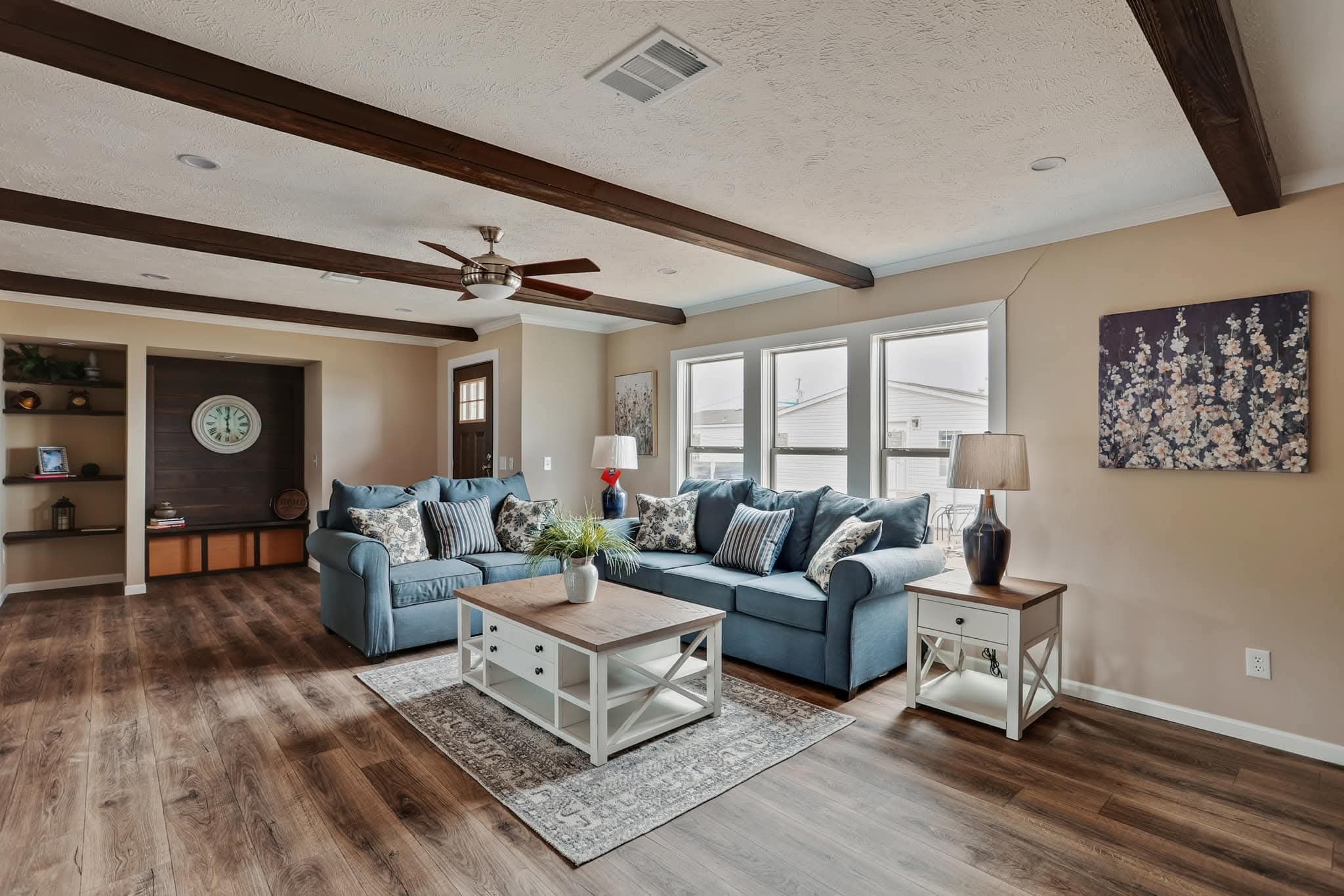 A cozy living room with wooden floors and ceiling beams, featuring two blue sofas with patterned cushions, a rug, and a coffee table. Light streams in through large windows, and artwork decorates the walls, creating a warm, inviting atmosphere.